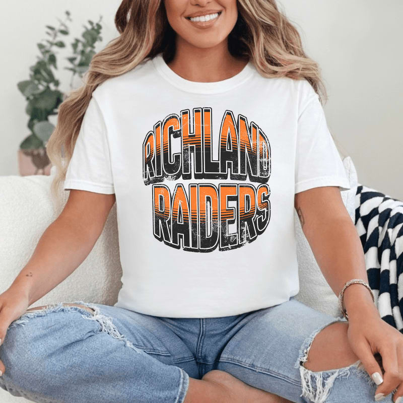 A woman in a white t-shirt with "RICHLAND RAIDERS" printed on it sits on a couch with a plant in the background.
