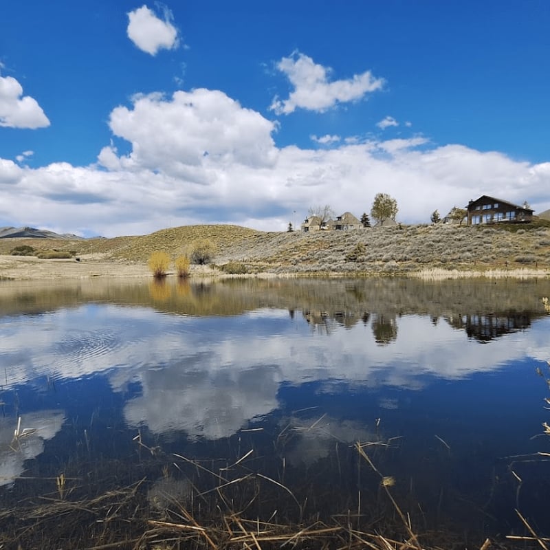 A calm lake in the foreground reflects the blue sky and fluffy clouds, while a cluster of houses sits on a grassy hill in the background.