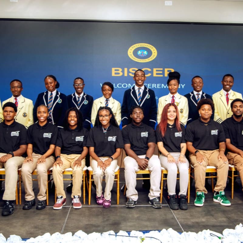 A group of students in uniform stand and sit in front of a backdrop with the school's logo and name.