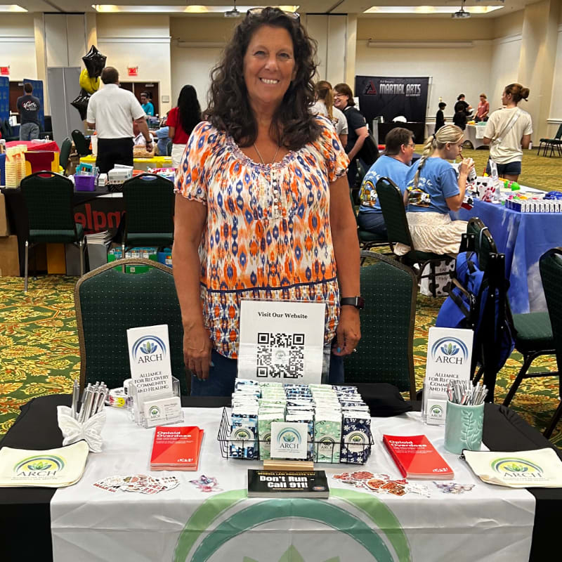 A woman stands behind a table displaying brochures and products at an indoor event, with other attendees and booths visible in the background.