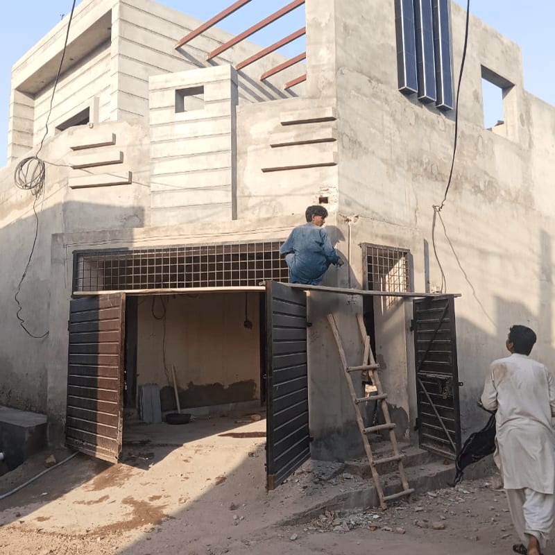 Two men work on the unfinished concrete facade of a modern house with a metal gate in the foreground and a clear sky in the background.