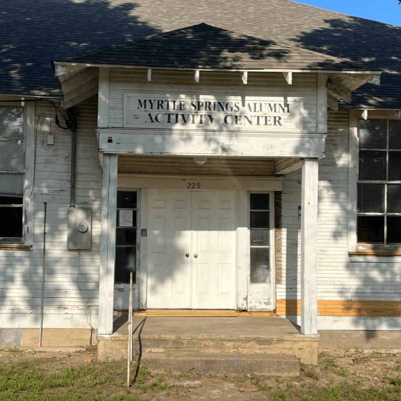 A weathered white building with "MYRTLE SPRINGS ALUMNI ACTIVITY CENTER" above double doors, set against a clear blue sky.