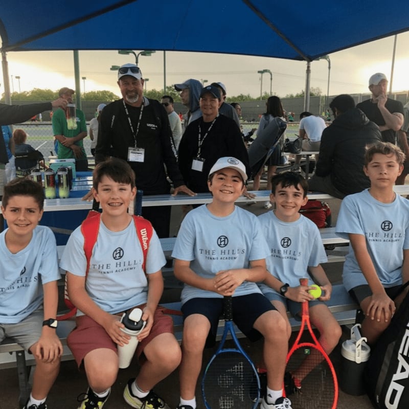 Six young boys in light blue shirts sit on a bench in the foreground with tennis rackets and bags, while adults and other children are visible in the blurred background under a blue canopy.