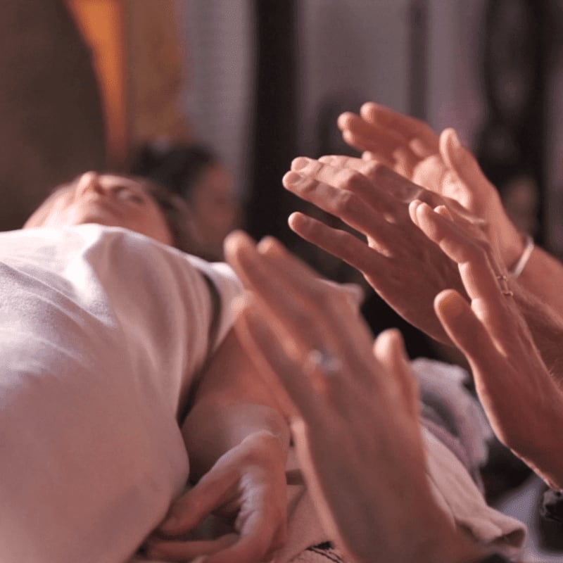 Hands are clapping in the foreground around a person lying down, with a blurred background of people and warm lighting.