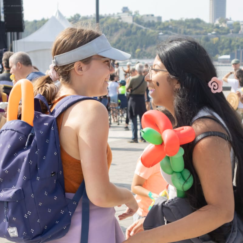 Two young women with a balloon animal smile at each other in the foreground, while a blurred crowd and tents are visible in the background.