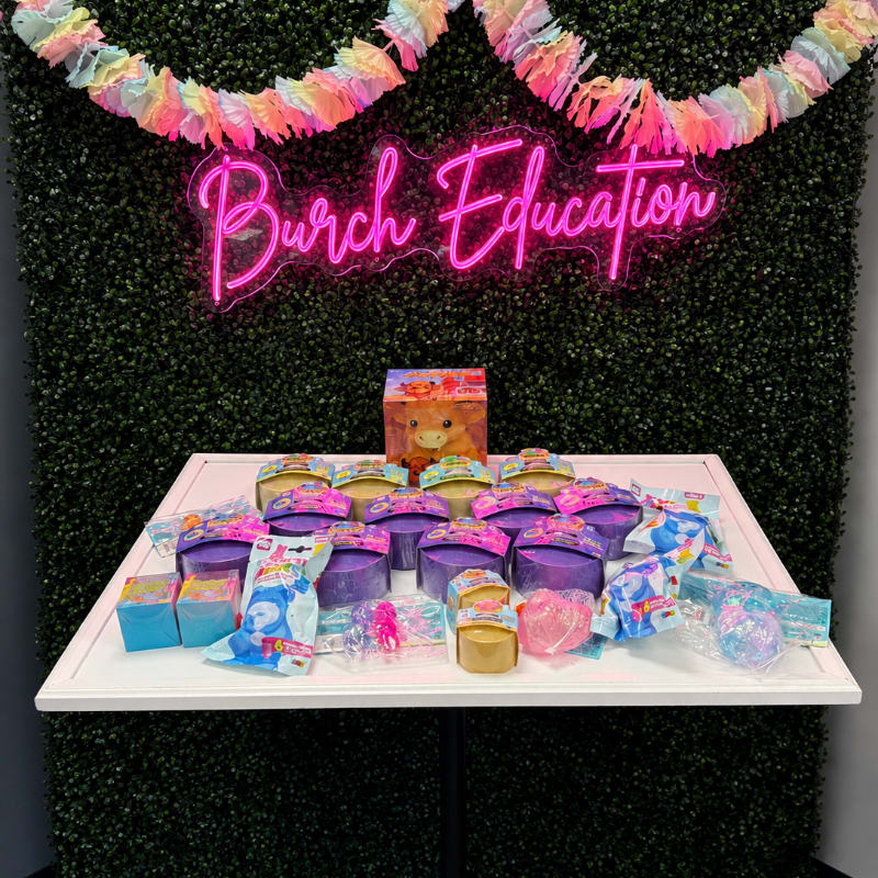 A white table laden with colorful toys sits in front of a lush green hedge adorned with a bright pink "Burch Education" neon sign and a festive pastel garland.