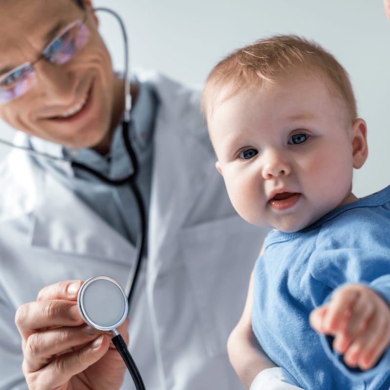 A smiling doctor in a white coat holds a stethoscope to a baby in a blue onesie, with a blurred background.