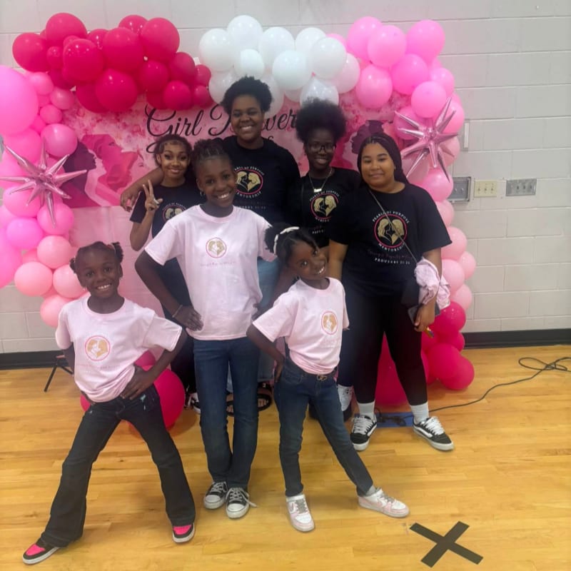 A group of young girls and women pose in front of a pink and white balloon arch and a "Girl Power" banner, with a wooden floor and a black "X" marking in the foreground.