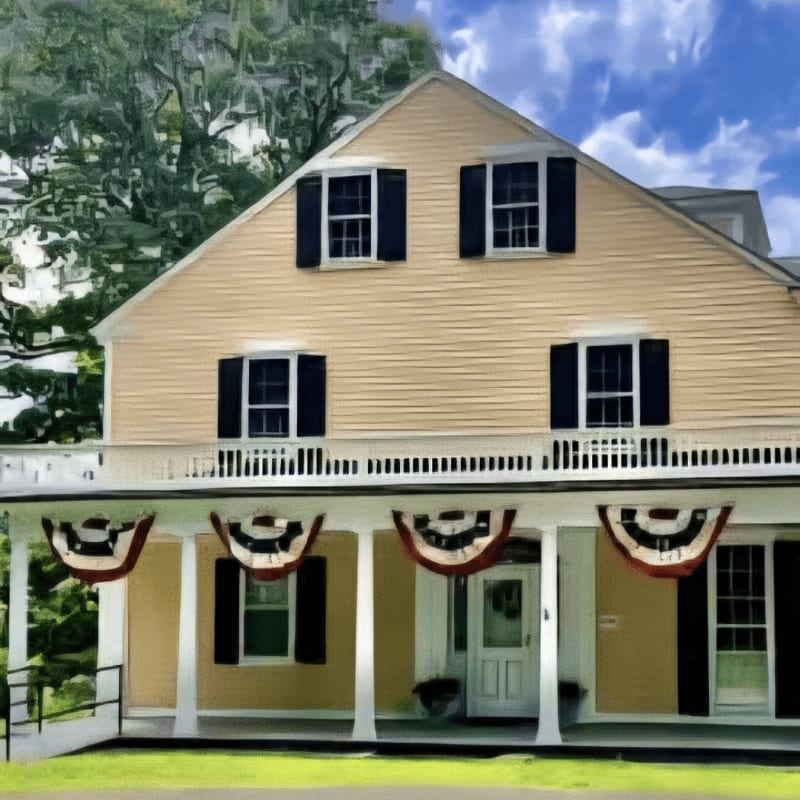 A light yellow house with black shutters and patriotic bunting on its porch stands against a backdrop of green trees and a blue sky.