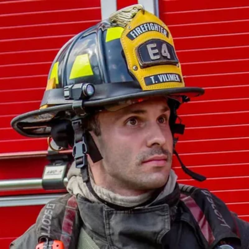 A firefighter in a helmet and uniform stands in front of a red, textured background.
