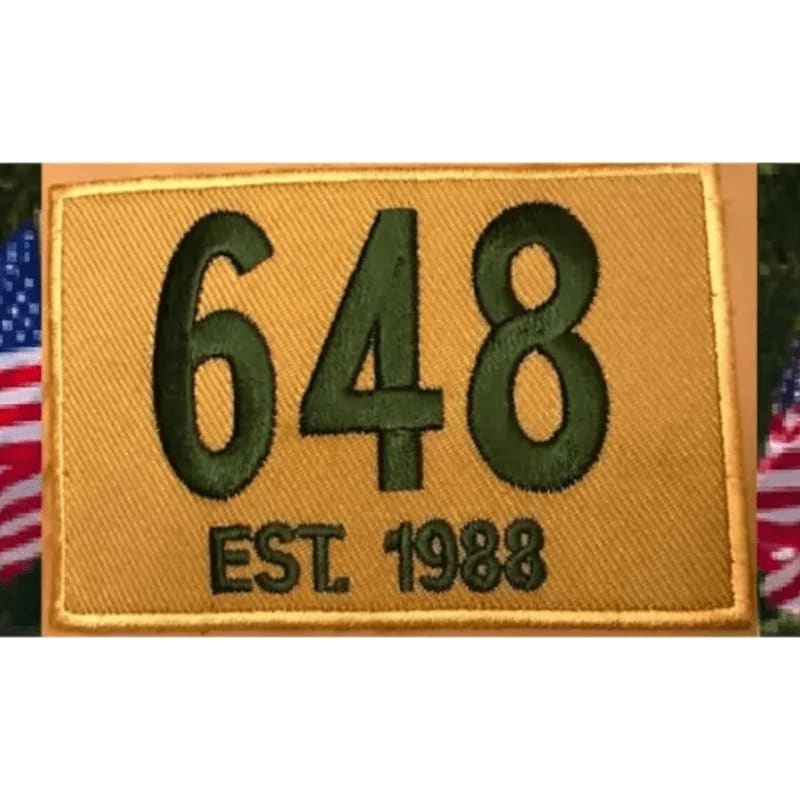 A gold embroidered patch with "648 EST. 1988" in green lettering is framed by American flags.