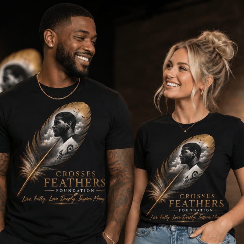A smiling Black man and a smiling blonde woman stand side-by-side, both wearing black t-shirts featuring a feather graphic with a portrait and text, against a dark, softly lit background.