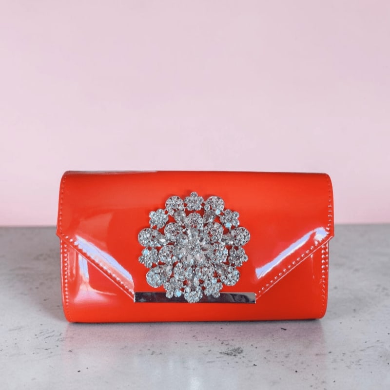 A shiny orange clutch adorned with a sparkling brooch sits on a textured surface against a soft pink background.