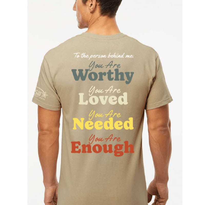 A man wearing a tan t-shirt with empowering messages printed on the back stands against a white background.