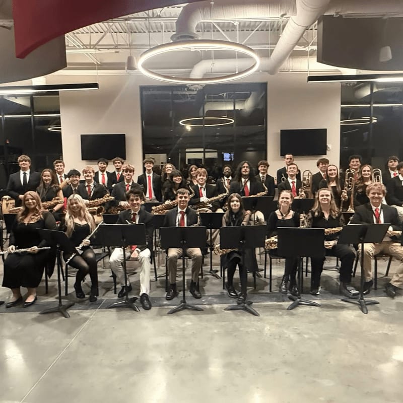 A group of young musicians in formal attire are seated in a modern room with large windows and contemporary lighting.
