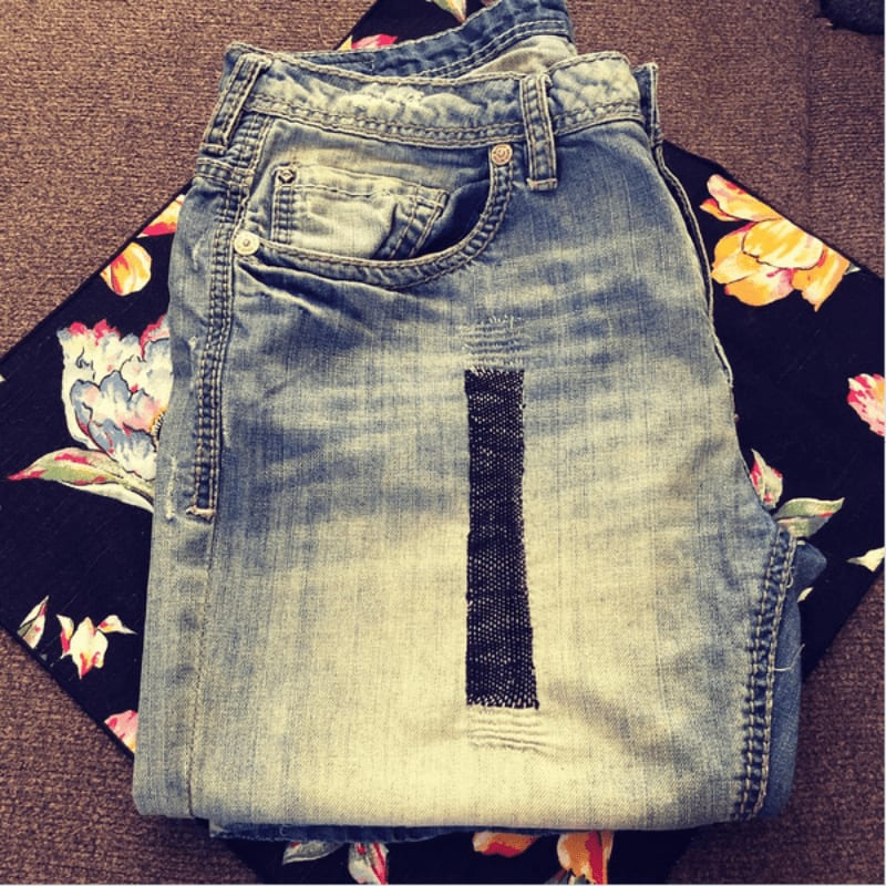 A pair of distressed denim jeans with a patch lies on a black fabric with a floral pattern.