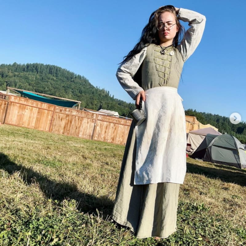 A young woman in historical attire stands in a grassy field with tents and a wooden fence in the background under a clear blue sky.