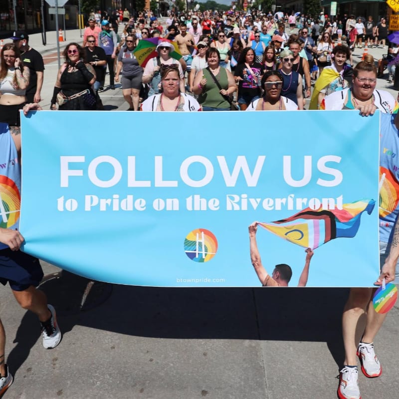 A large group of people march in a parade, holding a banner that says "FOLLOW US to Pride on the Riverfront" with a crowd of onlookers in the background.