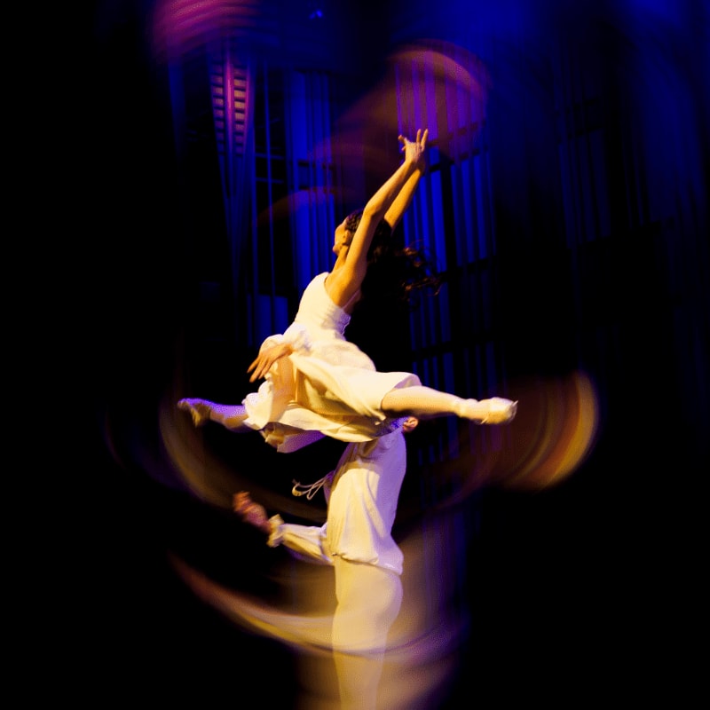 A dancer in a white dress is lifted by a partner against a blurred, colorful background of blue and purple lights.