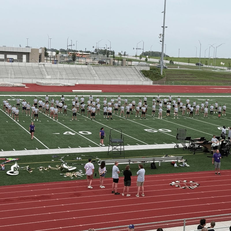 A marching band practices on a green field in front of stadium seating, with a road and buildings visible in the background.