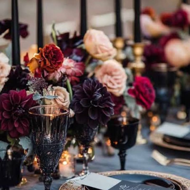 A dark, moody floral arrangement with deep red and purple blooms sits on a table set with black goblets and gold-rimmed plates, with tall black candles in the background.
