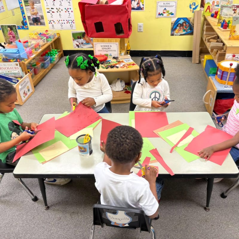 Four young children are seated at a table cutting red and green paper in a classroom filled with toys and educational materials.