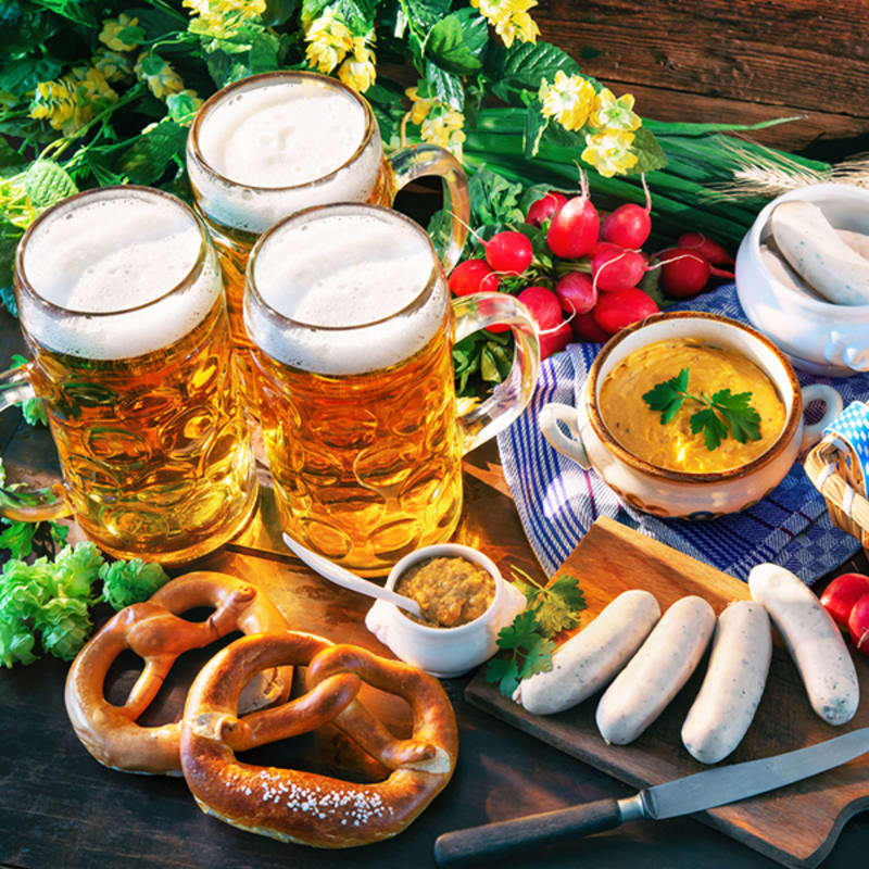 Three frothy mugs of beer sit on a wooden surface with pretzels and mustard in the foreground, while radishes, sausages, and greenery fill the background.