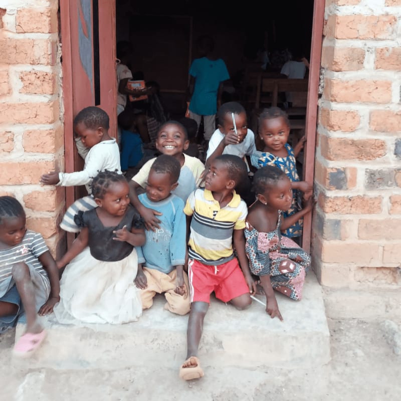 A group of smiling children are gathered in a doorway, with a classroom visible in the background.