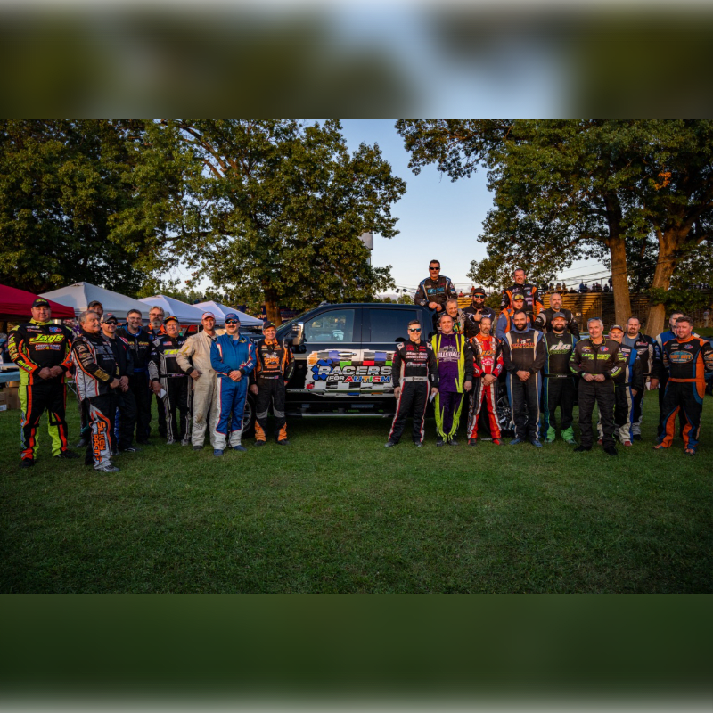 A group of people in racing suits stand in front of a van decorated with stickers, with trees and a clear sky in the background.