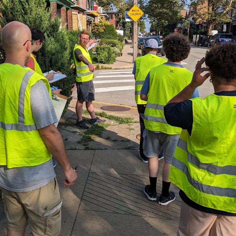 Several people in bright yellow safety vests stand on a sidewalk in the foreground, while a street with cars and houses is visible in the background.