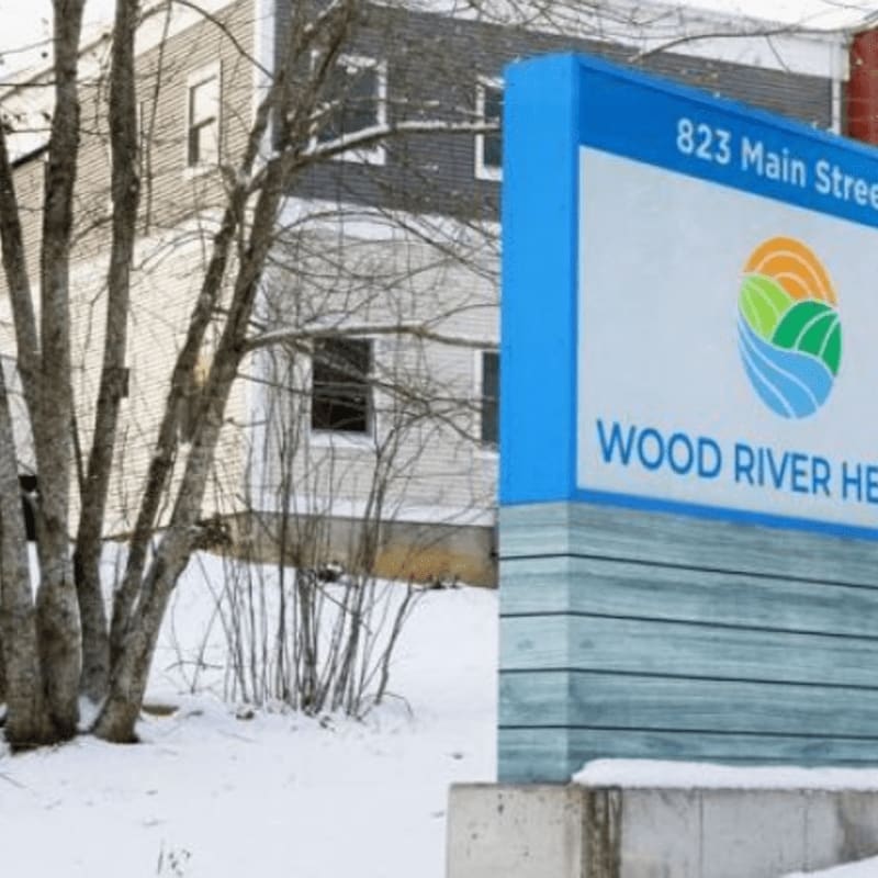 A blue and white sign for Wood River Health stands in the foreground against a backdrop of a snow-covered building and bare trees.