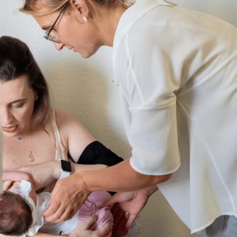 A woman in the foreground is breastfeeding a baby, while another woman in the background leans in to assist.