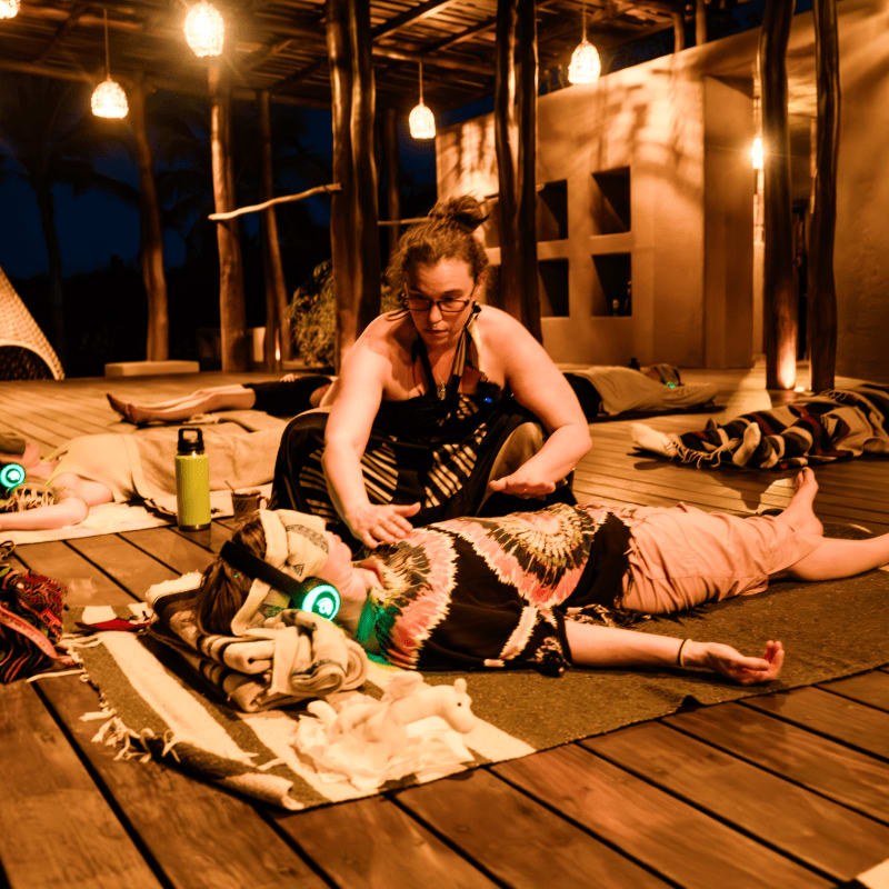 A woman in the foreground receives a healing session from another woman, while others lie on mats in the dimly lit, open-air background.