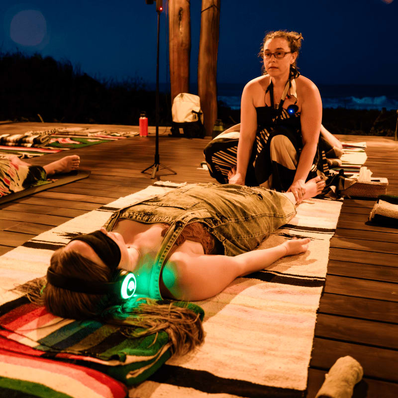 A woman lies on a striped blanket on a wooden deck with glowing headphones, while another woman kneels beside her, offering a massage, with a dark, twilight sky in the background.