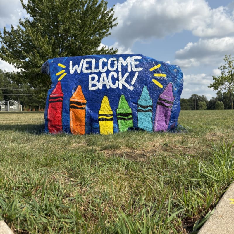 A large blue rock painted with "WELCOME BACK!" and colorful crayons sits in a grassy field with trees and a building in the background.