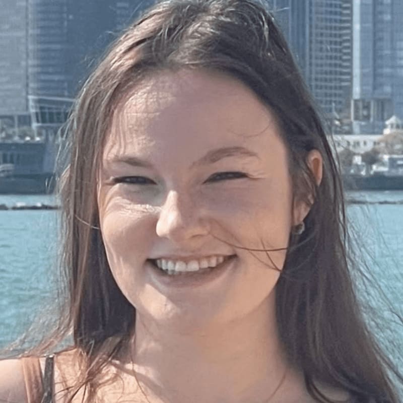 A young woman with a wide smile is in the foreground, with a blurred cityscape and water in the background.