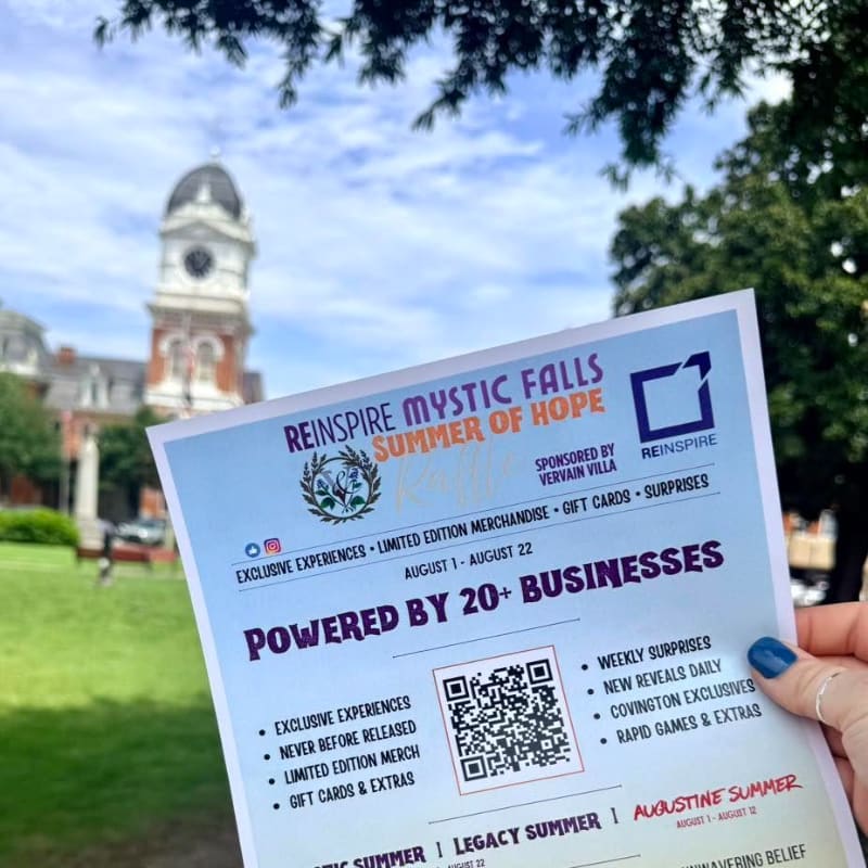 A flyer for "Reinspire Mystic Falls Summer of Hope" is held in the foreground, with a clock tower and building in the blurred background.