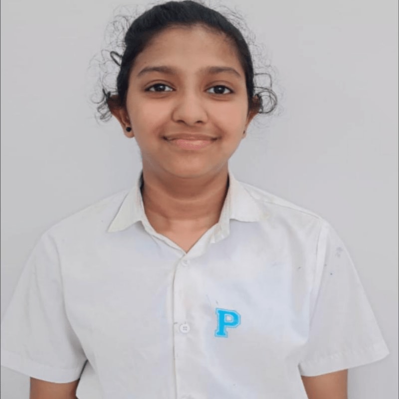 A young girl with dark curly hair pulled back smiles at the camera, wearing a white collared shirt with a blue "P" logo, against a plain white background.