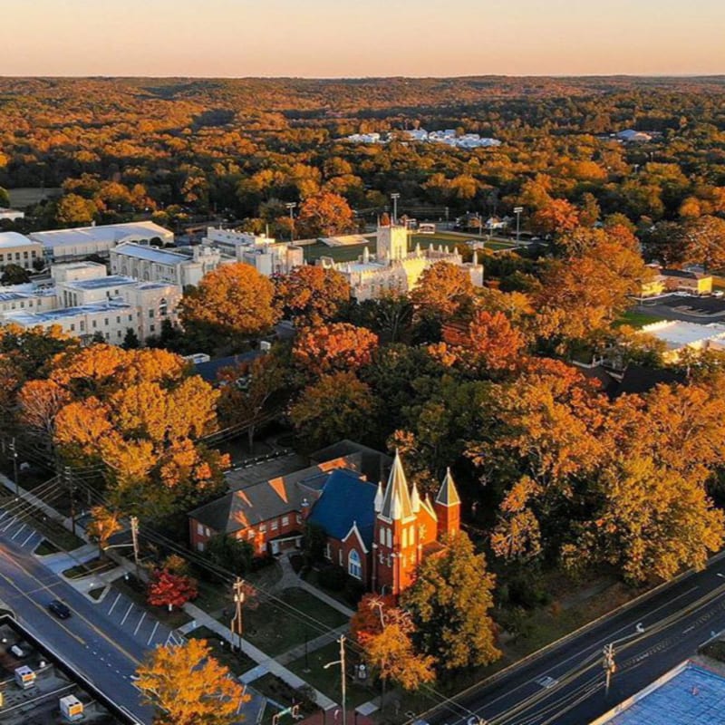 A red brick church with a blue roof sits in the foreground, surrounded by trees ablaze with autumn colors, while a sprawling white campus complex is visible in the background under a warm, hazy sky.
