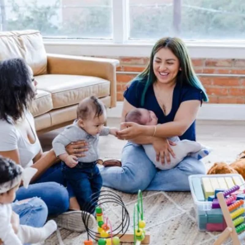 Two women are sitting on the floor with babies in a room with a couch and toys.