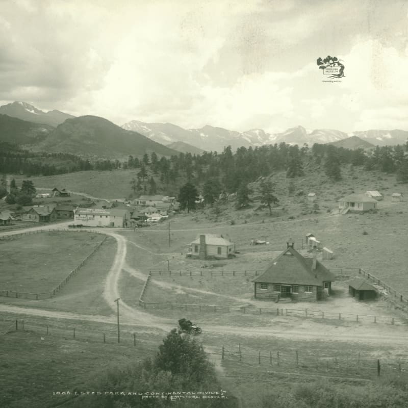 A dirt road winds through a rural landscape with a few buildings in the foreground, leading to a small town nestled in a valley with a majestic mountain range in the background.