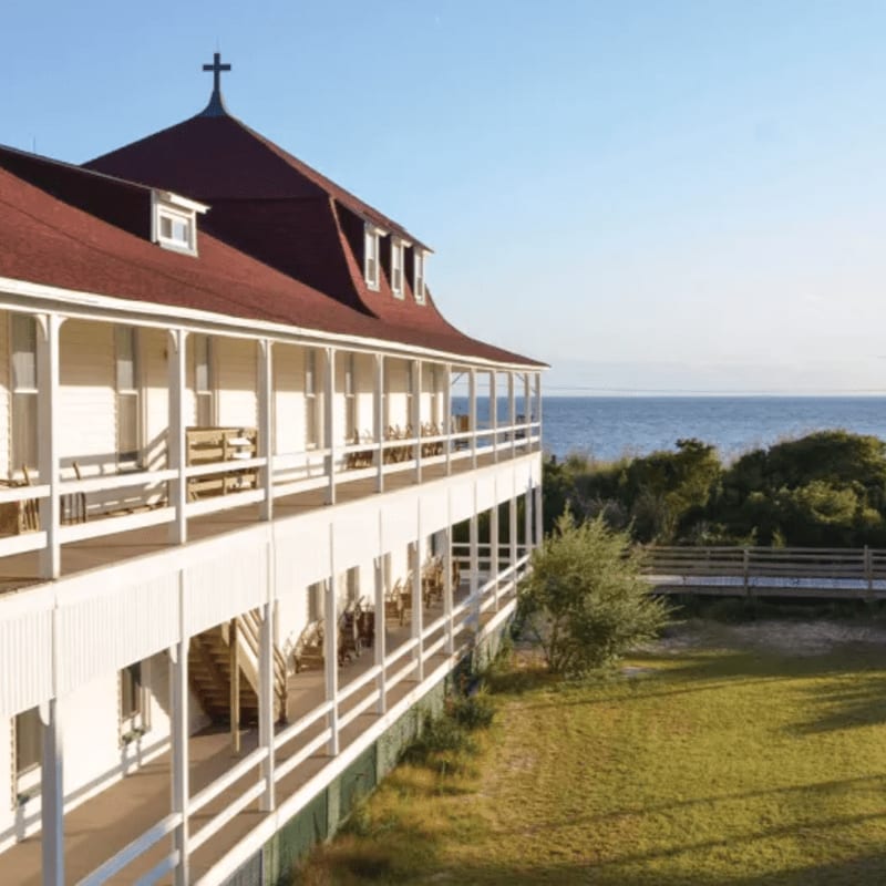 A white building with a red roof and a long balcony overlooks a grassy lawn and the ocean.
