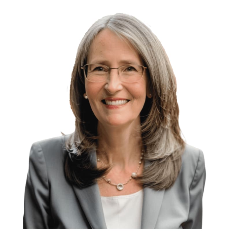 A smiling woman with graying hair and glasses, wearing a gray suit and pearl necklace, is set against a plain white background.