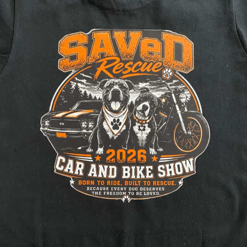 Two dogs wearing bandanas sit in front of a classic car and a motorcycle, with "Saved Rescue 2026 Car and Bike Show" text above and below, set against a backdrop of trees and a cloudy sky.
