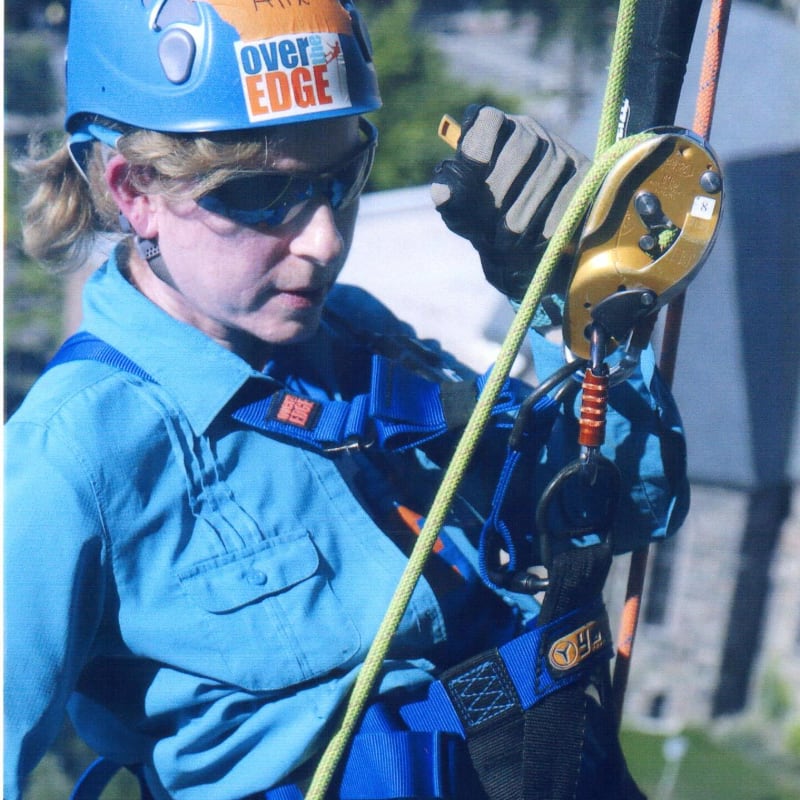 A woman in a blue shirt and harness is rappelling down a building, with a blurry cityscape in the background.