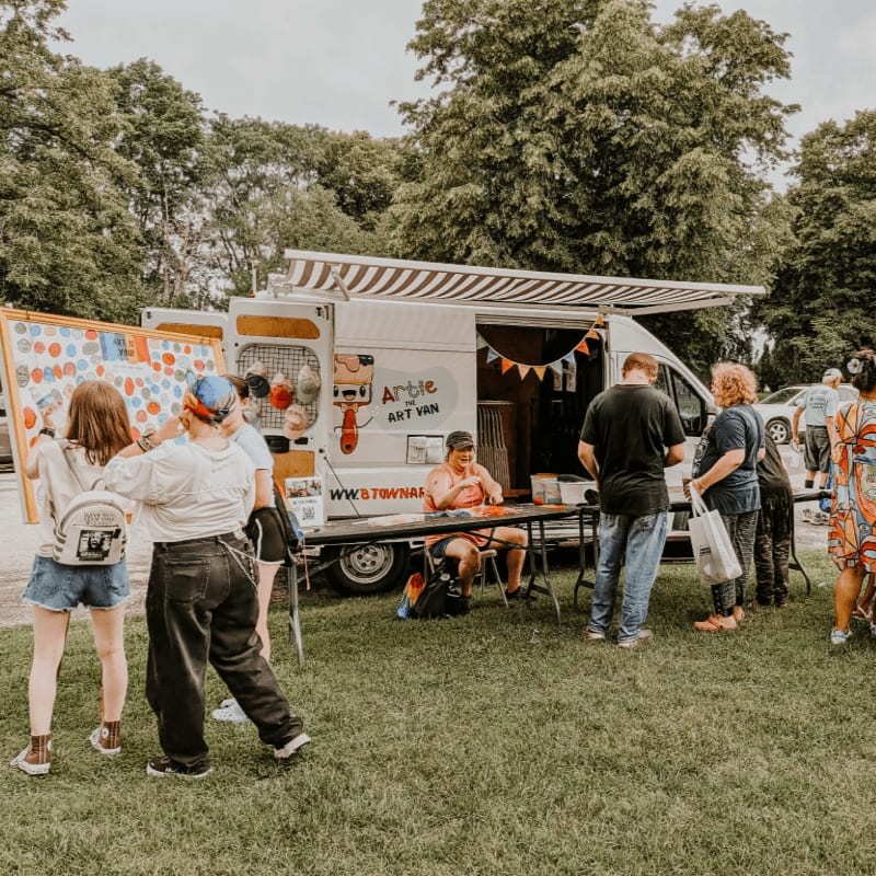 People gather around an art van with a colorful display board in the foreground, set against a backdrop of trees and a cloudy sky.