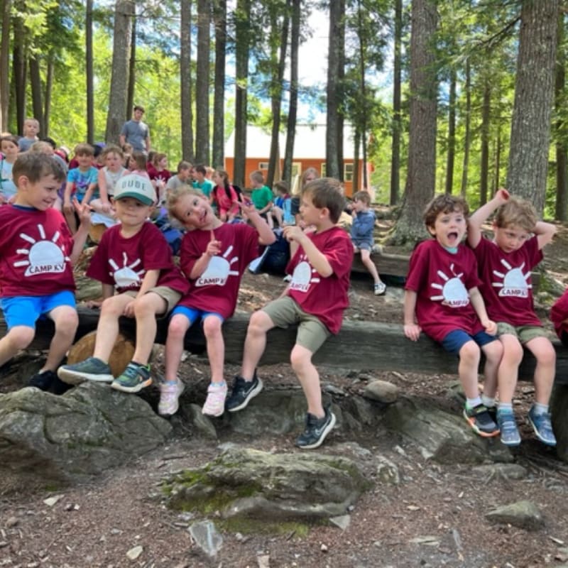 Children in maroon shirts sit on a log in the foreground, with a wooden building and trees in the background.