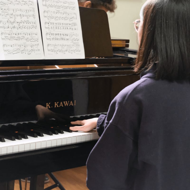A young woman plays a Kawai piano with sheet music in front of her, while another person's reflection is visible in the polished wood.