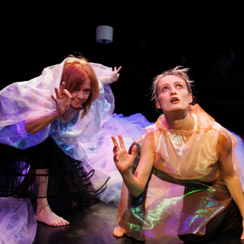 Two women in translucent, iridescent costumes are on a dark stage, with one on the left reaching forward with claw-like hands and the other on the right looking upwards with an open mouth.