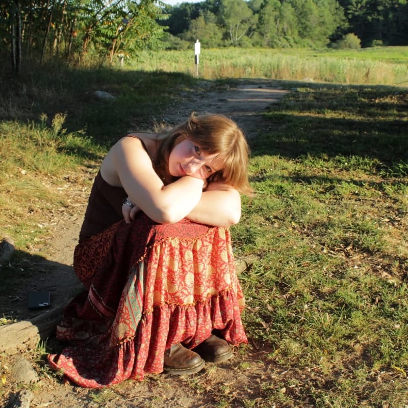 A young woman in a long, patterned skirt sits with her knees pulled to her chest on a grassy path, with a sunlit field and trees in the background.
